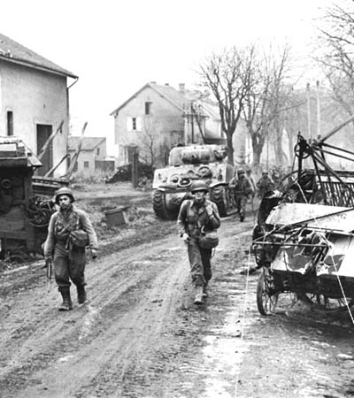 Photograph: Entering The Outskirts of Metz, men of the 378th Infantry are shown on the morning of 17 November in pursuit of the enemy along roads strewn with abandoned equipment.